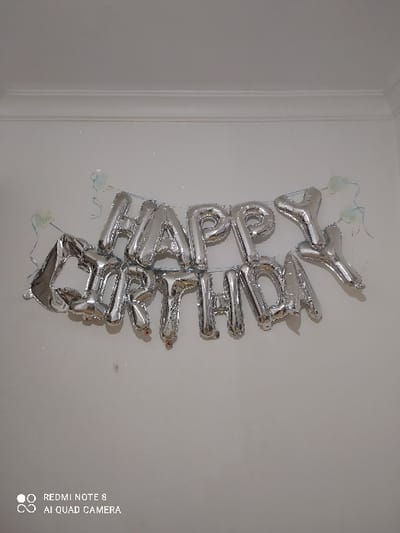 Silver "Happy Birthday" balloons are mounted on a white wall, arranged vertically. The letters are inflated and have a metallic shine. Thin, clear strings with remnants of adhesive hold the balloons in place. The wall appears clean and slightly textured. A corner of a door frame is visible on the left. The image is shot in portrait orientation.
