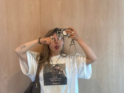 A woman with long brown hair poses for a selfie in front of a light brown wood panel wall. She wears glasses, a white t-shirt with a graphic, a silver necklace, and a bracelet. She holds her phone in both hands, with a ring on her right hand. The phone has a wrist strap. She sticks out her tongue and makes a peace sign near her face. A tattoo is visible on her left arm. A dark bag hangs from her shoulder.