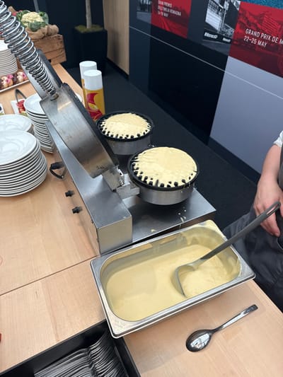 The image depicts a food service station, specifically a waffle preparation area, located within a hospitality or catering zone at the Circuit de Spa-Francorchamps in Stavelot, Belgium.
In the foreground, a light-colored wooden countertop features a stainless steel commercial waffle maker with two circular cooking molds, both containing uncooked, light-yellow waffle batter that slightly overflows. To the right of the waffle maker, a stainless steel GN 1/3 pan, marked "GN 1/3 111LAH," is filled with additional waffle batter, holding a dark grey serving ladle. A single silver spoon lies on the countertop to the right of the batter pan.
On the left side of the countertop, a tall stack of clean white ceramic plates is present. Adjacent to this, a black tray containing multiple silver forks is visible. Further left, a wooden crate holds green vegetables, possibly cauliflower or broccoli, and a tray with small, dark pastries or donuts. Two white spray cans with yellow accents, one featuring a red symbol, are positioned behind the waffle maker.
The background consists of a dark grey wall adorned with large promotional posters for motorsport events. Clearly visible are posters for "GRAND PRIX DE MONACO 22-25 MAY" and "GRAND PREMIO DELL'EMILIA ROMAGNA 18-18 MAY," indicating a motorsport-related venue. A tall, dark planter with a green plant is situated in the left background. On the far right, the arm and torso of a person, wearing a dark grey apron over a white shirt, are partially visible, suggesting a server or attendee. The scene indicates an active food preparation and serving setup.