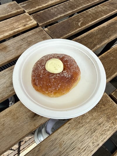 A single, sugar-coated donut with a light-yellow dollop of cream or butter centered on its top is positioned on a circular, white, disposable plate. The plate rests on a weathered wooden slatted table, characterized by visible grain and minor surface wear. Below the table, a dark ground surface is partially visible through the gaps between the wooden slats, along with a partial view of a person's foot wearing a light-colored shoe and a grey sock. The image lacks specific visual identifiers to determine a precise location within Chula Vista, United States of America.