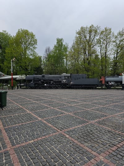 The image depicts a steam locomotive on display in Arkhangel’skoye, Russia. The locomotive is black and sits on a paved area with grey and red bricks arranged in a grid pattern. In the background, there are green trees and an overcast sky. A portion of a vintage airplane can be seen near the locomotive.