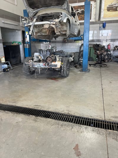 A car body is lifted by a blue hydraulic lift in a garage setting. Beside it, the car's chassis with wheels is present, suggesting disassembly. Scattered car parts and tools fill the background, and a floor drain runs along the edge of the concrete floor, indicating a repair facility. The overall scene suggests a complex automotive repair or restoration project is underway.