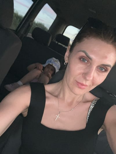 The image is a selfie taken inside a vehicle. A woman with blonde hair and blue eyes is in the foreground, looking at the camera and smiling. She is wearing a black top with a necklace and a cross pendant. In the background, a child is lying down, wearing a blue and white hat. The vehicle appears to be moving through a rural area, with trees visible outside the windows. The lighting suggests it is either dawn or dusk. There are no visible landmarks or elements that place this scene specifically in Aqsu city administration, Kazakhstan.