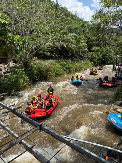 The image shows a whitewater rafting scene on a river in Tambon Song Phraek, Thailand. Multiple inflatable rafts filled with people are navigating through rapids. The rafts are of different colors including red, blue, yellow, and black. The people in the rafts are wearing life jackets and using paddles to steer. The river is surrounded by lush green vegetation, including trees and bushes. A metal bridge railing is visible in the foreground. The water is choppy and brown, indicating fast-flowing currents.
