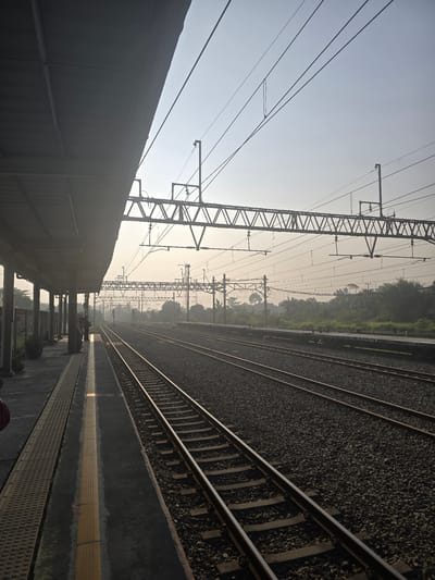 This is a photograph of a train station platform in Klapanunggal, Indonesia, taken during the day. The image shows multiple sets of train tracks running parallel to the platform. The platform itself is covered by a large, dark gray canopy supported by pillars. A yellow line runs along the edge of the platform. In the background, there are overhead electrical wires and structures for the train system, as well as lush green vegetation and trees. The sky is hazy and appears to be either early morning or late afternoon due to the soft lighting. A few people are visible standing on the platform, appearing to wait. The overall scene depicts a functional and active train station environment.