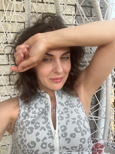 A woman lies on her back on a white woven chair. Her left arm is bent and resting on her forehead, with her hand covering part of her face. Her right arm is extended upwards, out of frame. She wears a grey and white leopard-print garment that appears to be a romper or jumpsuit, zipped down the front, revealing a silver cross necklace. Her dark hair is spread out around her head, and she is looking at the camera with a slight smile. The background is blurred, but appears to be outdoors with sunlight suggesting daytime. The texture of the woven chair is prominent in the foreground.