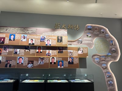 A display wall features a grid of portraits with accompanying text, likely honoring individuals. The portraits are arranged in three rows, with a dark-toned background and clear cases displaying books or documents below. To the right, a stylized artwork depicts a tree-like shape with circular images and text embedded within, potentially representing growth or connections. The wall is painted in shades of gray and tan.
