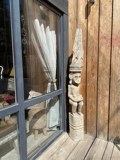 A carved wooden sculpture rests on the gray-framed window of a shop in Askat, Russia. The sculpture is light in color and appears to depict a human figure with intricate designs at both ends. The window reflects the wooden planks of the building's exterior. Inside the shop, several items are visible, including a white curtain, a woven basket stool, decorative stones, and fabrics of various patterns.