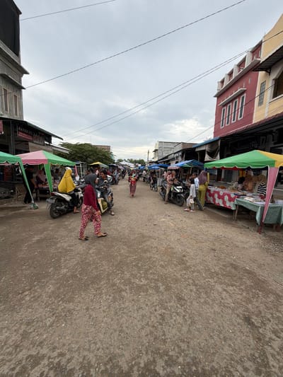 The image depicts an active street market in Ietarek Dua, Indonesia, under an overcast sky. The unpaved dirt and gravel road serves as the primary thoroughfare, lined on both sides by a mix of permanent buildings and temporary market stalls.

On the left, a tall, multi-story building with a grey, unfinished concrete facade is visible, beneath which a shop displaying an "ASPIRA OIL" sign operates. Adjacent to it are multiple market stalls featuring green and pink striped awnings, where individuals are visible, including a person wearing a yellow hijab near a parked white scooter. Further right on the left side, another individual on a black motorcycle is positioned near a person in a dark red shirt and pink patterned pants, who is walking towards the right side of the frame.

The right side of the street features a distinct multi-story building with a red facade and a yellow upper section, characterized by several elongated white window panels. In front of this building, several market stalls with green and yellow striped awnings display various goods on tables, including packaged items and textiles. Individuals, including a person in a light green top and a child, are seen near these stalls, suggesting vendor or shopper activity.

The midground and background show the street extending into the distance, populated by numerous pedestrians and motorcycles. More market stalls and buildings line the road further down, with trees visible in the far background. Overhead utility lines cross the sky. Multiple motorcycles are parked or moving slowly throughout the scene, indicating a bustling commercial environment.