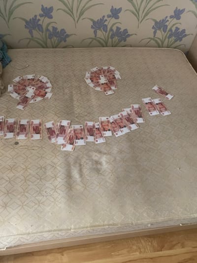 The image depicts a mattress on the floor, covered with Russian Rubles arranged to form a smiley face. The currency appears to be 5000 Ruble notes. There are two circular arrangements of bills at the top, resembling eyes, and a line of bills across the middle forming a mouth. A few scattered bills are also visible. The background shows wallpaper with a floral pattern. The setting is likely a domestic interior, possibly a bedroom, in Kovrov, Russia. There are no people visible in the image.