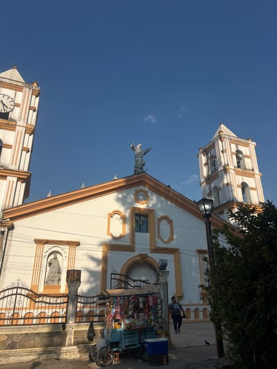 The image depicts a building, likely a church, with white walls and brown trim, featuring a tower with a clock face, a statue of Jesus Christ, and decorative elements. A street vendor with a cart selling snacks and drinks is positioned in front of the building, and a person stands nearby. The photo appears to be taken in La Esperanza, Honduras, based on the architecture and Google Lens search analysis. The sky is blue, suggesting a sunny day.