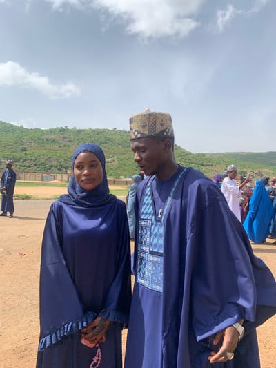 The image shows a man and a woman posing outdoors. The man wears a dark blue robe with patterned embroidery and a patterned kufi hat. He has a watch on his left wrist. The woman is wearing a dark blue hijab and robe. Behind them is a green hillside and a cloudy sky. Other people are visible in the background. They are located in the city of Naraguta Hausawa, Nigeria, as indicated by the location data.