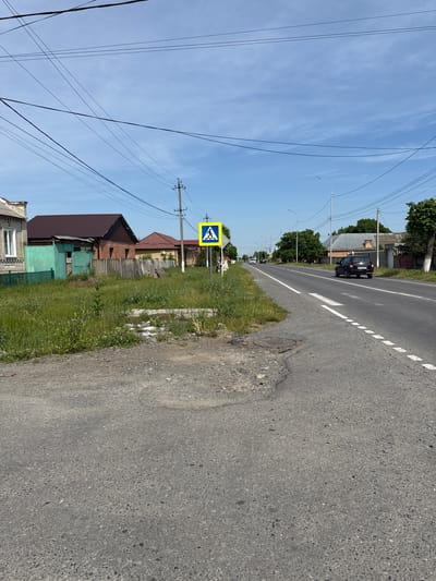 The image shows a rural road in Nogir, Russia, on a clear, sunny day. The road is paved and has white lane markings. On the side of the road, there is a pedestrian crossing sign, indicating a safe place for people to cross. A black car is driving on the road. There are houses and trees visible in the background, suggesting a residential area. Overhead, power lines crisscross the sky, a common sight in many rural and suburban areas. The overall scene depicts a quiet, everyday moment on a Russian road. There is no visible text in the image.