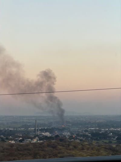 A landscape view with smoke rising from a distant fire in a town or city. The sky is hazy, transitioning from light to a slight orange hue. There are industrial chimneys in the middle ground, and houses with trees on either side. A vertical black wire cuts through the foreground, adding a sense of depth to the scene. The composition is slightly off-kilter.