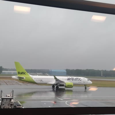 An AirBaltic Airbus A220-300 aircraft, bearing the registration YL-CSF, stands on a wet asphalt tarmac under an overcast sky at an airport in Skulte, Latvia. The aircraft features a white fuselage with "airBaltic" branding in black, a lime green tail fin with "Baltic" in black, and lime green engine nacelles also displaying "airBaltic." Reflections of interior rectangular light sources from the viewing window are visible on the upper part of the image and on the wet tarmac surface. In the background, a dense treeline borders the airfield, behind which are several airport structures: a long, low white building to the left, and a larger, light blue hangar displaying faint "AIRPORT BALTIC" text in the center-right. Several smaller, white aircraft are parked near the treeline. Airport ground equipment, specifically the upper portion of a mobile passenger stair or baggage conveyor, is partially visible in the bottom-left foreground. The image is framed by a dark horizontal window sill at the bottom.