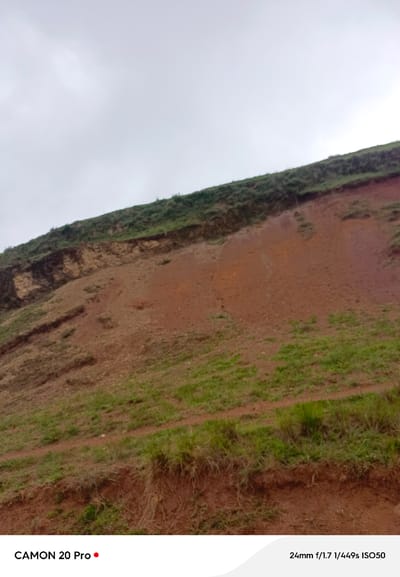 The image shows a steep, eroded hillside in Nguroje, Nigeria, with reddish-brown soil and patches of green grass. The sky is overcast and a dirt path winds along the bottom of the hill. Text in the lower left corner reads "CAMON 20 Pro" and in the lower right corner reads "24mm f/1.7 1/449s ISO50", indicating camera settings. There are no people or animals visible in the scene.