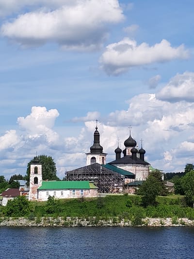 The image showcases a church complex situated in Goritsy, Russia, featuring a striking bell tower and a central church building adorned with black domes, all seemingly undergoing some form of construction or renovation, as evidenced by the scaffolding. The architectural style is distinctly Russian Orthodox. The buildings are positioned on a slightly elevated embankment bordering a body of water. Lush green vegetation surrounds the structures, while the sky is filled with puffy, white clouds against a blue backdrop. The water surface is dark, reflecting the sky above. The overall scene is serene and suggests a site of historical or religious significance.