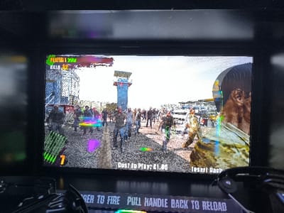 The image displays an arcade game screen, showing a first-person shooter with a zombie theme. The game scene depicts numerous grotesque zombie figures advancing towards the player across an open, desolate landscape featuring industrial-style structures, possibly watchtowers and fences, under a bright, clear sky.

On the game screen, user interface elements are visible: "PLAYER 1" with a health bar, "KILLS: 32", and a visible ammo count of '7'. Colorful visual effects, such as glowing outlines and streaks, are seen around some of the zombies, suggesting gameplay interactions. The bottom of the screen indicates "Cost to Play: £1.00" and "Insert Cash", signifying the game is ready for payment.

Below the screen, a portion of the arcade machine's control panel is visible, including part of what appears to be a light gun or rifle controller. Text on the control panel provides instructions: "TRIGGER TO FIRE PULL HANDLE BACK TO RELOAD". The activity taking place is someone engaging with, or preparing to play, this arcade game.

The media file captures a moment of arcade gaming in Agios Dimitrios, Greece. The time of day in the game appears to be bright daytime.