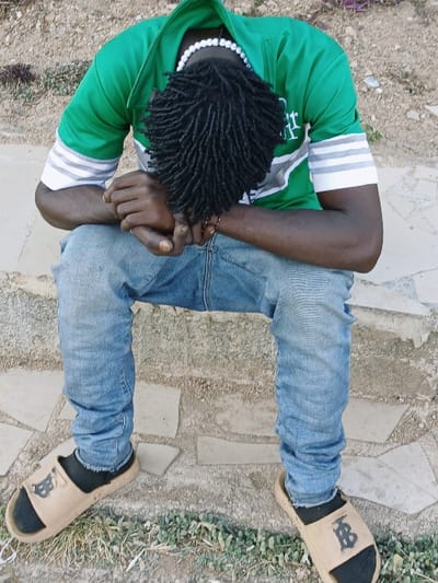 The image displays a person, presumably male, seen from a high-angle perspective, rotated 90 degrees counter-clockwise, appearing to be sitting or squatting on a textured outdoor surface. The person has dark skin and black dreadlocks, which are visible from the back of the head.
They are wearing a bright green hooded jacket or hoodie with prominent white and grey striped bands on both upper arms, consisting of two white stripes flanking a central grey stripe. A white beaded necklace is visible around their neck. The person is also wearing light blue denim jeans with a slightly faded appearance and some wrinkling, suggesting a relaxed fit.
On their feet, they are wearing black socks with light brown or tan slide sandals. The sandals each feature a dark, stylized letter 'B' or a similar emblem on the strap. The person's arms are bent at the elbows, with their hands clasped together or resting on their lap/knees.
The background consists of an uneven, earthy ground covered with various scattered pieces of light-colored, possibly concrete or stone, fragments that form an irregular pattern. There are also patches of small green plants or weeds visible on the right side of the image. The overall setting appears to be an outdoor, somewhat unpaved area. The lighting suggests it is daytime.