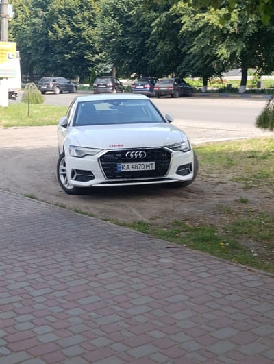 The image shows a white Audi A6 parked on a dirt road, with a sidewalk made of red and gray pavers in the foreground. The car is the primary subject, with its front grille and license plate clearly visible. The license plate reads "KA 4870 MT". Above the license plate, the Audi logo is prominent, and a red "CLAAS" logo is visible on the hood.
In the background, several other cars are parked along a street lined with mature trees, suggesting a residential or commercial area. The trees are lush and green, indicating it's likely daytime during the warmer months. The sky is not visible, but the lighting suggests it's a bright, possibly sunny day. The scene appears to be in Khorol, Ukraine. There are no people visible in the image, and the activity is simply the car being parked. The overall impression is a straightforward, static shot of a car in its environment.