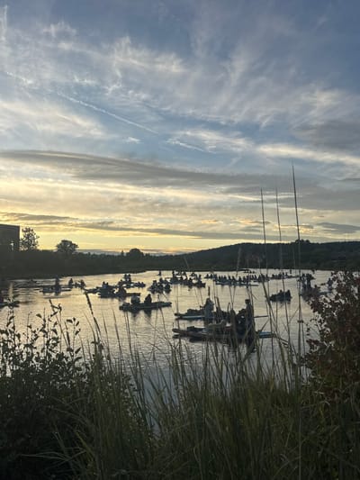 The image displays a crowded recreational scene on the Deschutes River in Bend, United States, at dusk. A low sun, positioned near the horizon, casts a warm, golden reflection across the water, silhouetting the distant landscape and the subjects within the frame. The sky above is characterized by layered cloud formations, including wispy cirrus and broader stratocumulus, with multiple linear contrails distinctly visible.
The river is densely populated with an estimated 50 to 70 individuals distributed across various personal watercraft. These vessels primarily consist of stand-up paddleboards, numerous inflatable rafts, and other small boats. Individuals are observed engaged in leisure activities, either paddling their craft or drifting stationary on the water.
In the immediate foreground, out-of-focus tall grasses and riparian foliage are present, suggesting the vantage point is from the riverbank. The background features a silhouetted skyline: buildings and trees are discernible on the left riverbank, while a tree-covered hillside occupies the right.
