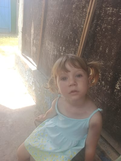 A young girl with blonde pigtails is sitting on a concrete surface in front of a dark, weathered wooden wall. She is wearing a light blue dress with a yellow lemon pattern. Sunlight is casting a bright glare on the left side of the image, illuminating the wall and a peeling paint on a structure next to it. In the background on the far left, a light blue wooden door is partially visible. The overall scene appears to be outdoors in a rustic or rural setting, potentially in Moldoveanca, Moldova, given the context. The girl is looking towards the camera with a neutral expression.