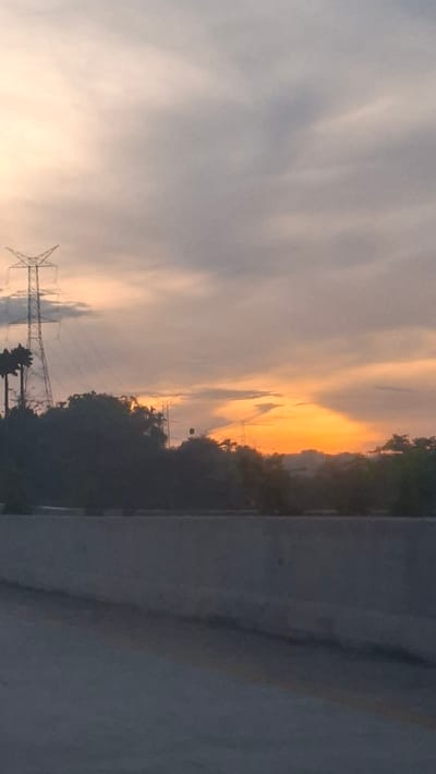 The image captures a sunset view from a highway overpass in Lonbaroh, Indonesia. The sky is filled with soft, diffused light, with patches of orange and pink near the horizon, indicating the sun is setting or has recently set. Wispy clouds drift across the sky. In the middle ground, dark silhouettes of trees and foliage obscure the landscape. A prominent power transmission tower stands tall on the left side of the frame, with its metallic structure visible against the sky. Another, smaller power line tower is faintly visible in the distance. A concrete barrier runs across the lower part of the image, suggesting the perspective is from a raised roadway. The overall scene conveys a sense of urban twilight encroaching on a natural and industrial landscape.