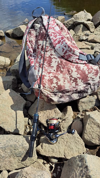 The image depicts a fishing rod and reel resting on large, light-colored rocks next to a body of water. A camouflage-patterned backpack, with shades of pink and beige, is situated behind the fishing gear. The backpack has gray straps and buckles. The fishing rod is black with a blue accent near the handle. The reel is silver and red with a spool of fishing line. The water appears calm, reflecting the sky. The scene is set outdoors, likely on the shore of a lake or river. The ground is covered with rocks of various sizes.