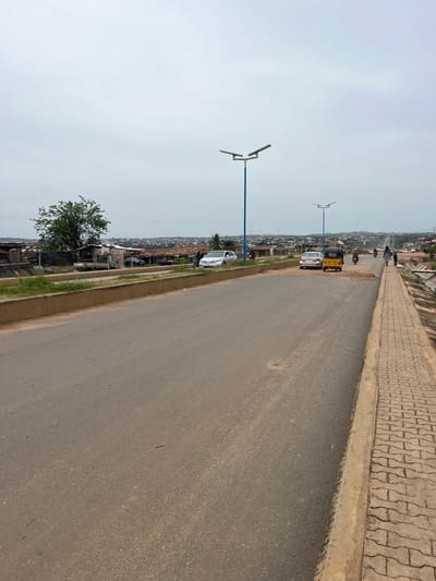 The image depicts a view of a street in Osogbo, Nigeria. The street is asphalt and has a light-colored sidewalk made of interlocking paving stones. A blue light pole stands on the left side of the road. The street has vehicles parked on the left-hand shoulder. A tree and several stalls or shops can be seen on the left, just off the road. In the distance, the road extends into the cityscape with houses and buildings visible. Some people are walking on the sidewalk in the distance. The sky is overcast.