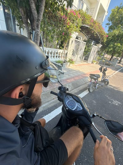 A first-person perspective shot shows a person wearing a black helmet and sunglasses, seated on a scooter. They have a beard and an earbud in their left ear. The person is wearing a dark blue long-sleeved shirt and black shorts. A black fanny pack is worn across their waist. The scooter's dashboard, handlebars, and mirror are visible in the foreground. In the background, a street is lined with trees and a white building with pink flowers growing on an overhead trellis. Two other scooters are parked on the side of the road. The scene appears to be in Cam Lâm, Vietnam, as indicated by a sign on a bench in the background that reads "CAM RANH".