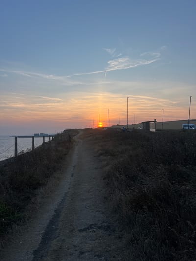 The image depicts a coastal path at sunset in Ovingdean, United Kingdom. The sun is low on the horizon, casting a warm glow across the sky and illuminating thin, wispy clouds. The sea is visible to the left, with a fence line running along the edge of the path. The path itself is dirt and winds uphill. On the right side of the path, there is dense, dry grass and a road with streetlights and a few vehicles, including a white van and a bus stop shelter. In the distance, there are some buildings, possibly residential, and a field behind the road. A single person is visible walking on the path. The overall scene is tranquil and evokes a sense of end of day.