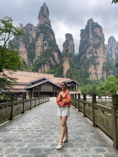 Here's a concise description of the image:

The image is a medium shot of a woman standing on a tiled pathway, with majestic rock formations visible in the background. The woman is wearing a patterned orange shirt and white shorts. To her left is a building displaying the UNESCO logo and Chinese text, potentially indicating a World Heritage Site. The pathway she's on is lined with carved wooden railings. The background features towering, mist-shrouded rock formations, typical of a mountainous region, suggesting a natural landmark location. The overall atmosphere is that of a tourist destination, with a blend of natural and man-made elements. Given the UNESCO logo, it's possibly a site of natural significance in the region.