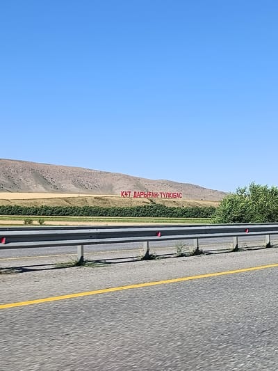 The image shows a vast, arid landscape under a clear blue sky. In the middle ground, large red letters are inscribed on a hillside, spelling out "ҚҰТ ДАРЫҒАН-ТҰЛКІБАС". This appears to be a landmark or signage in the area of Жас кешу, Kazakhstan. The letters are situated on a barren, rocky slope dotted with sparse vegetation. Below the hill, there is a strip of green foliage, possibly a field or a line of trees. In the foreground, a grey metal guardrail runs horizontally across the frame, separating the viewer's perspective from the road. The road itself is paved and has a bright yellow line marking its edge. The overall impression is one of a remote, open environment with distinct human-made signage.