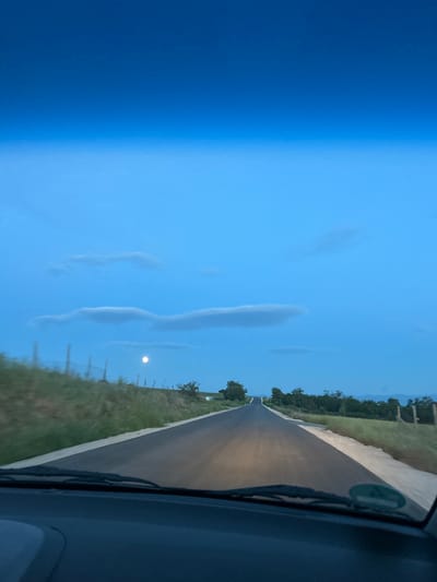 The image depicts a view from inside a vehicle, looking out onto a road. The road is asphalt, and there is vegetation on either side. In the distance, trees are visible. Above the road, the sky is light blue with elongated clouds. A small, bright circle is visible in the sky, possibly the moon or sun. The vehicle's windshield wipers are partially visible in the foreground. The location is unspecified, but the city of Smilets, Bulgaria is mentioned.