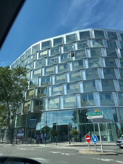 Here's a description of the image:
The image shows a modern, multi-story building with a distinctive facade composed of numerous square windows, angled slightly outward. The building has a curved architectural element. The sky is blue, with some reflections in the building's windows.
Located adjacent to the building is a street with traffic signs, including directions to Boulogne Centre (D50), Paris (Porte de St Cloud), and Quartier du Point du Jour. A "no entry" sign and an indication of a one-way street are also visible. People are standing on the sidewalk near what appears to be a bus stop with an informational sign and an advertisement. A tree is in front of the building. A car is parked on the right side of the image. The location is Boulogne-Billancourt, France, as indicated by the road sign to Boulogne Centre.