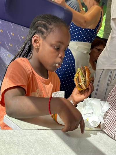 A young girl with braided hair and an orange t-shirt is holding a large hamburger. She is seated on a purple seat with a patterned backrest, and her arm rests on a textured surface. A woman in a blue and white polka dot dress is partially visible in the background, sitting in the same row. The scene appears to be indoors, possibly on public transportation, given the seating arrangement. The lighting suggests it could be daytime. The girl's expression is difficult to discern, but she is focused on the hamburger.