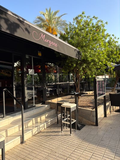 The image shows the outdoor seating area of a restaurant or cafe named "Morgana" in L'Alfàs del Pi, Spain. The establishment has a black awning with the name "Morgana" written in pink cursive. Large glass windows form the facade of the building, with the interior visible, showing tables and chairs. Outside, there are several tables and chairs set up on a paved patio. One table is a high-top with two bar stools. A menu board is positioned near some of the outdoor seating. The area is landscaped with gravel and a tree with green foliage and some red flowers. Palm trees are also visible in the background, suggesting a warm climate. The sky is clear and blue, and the sun is casting shadows, indicating it is daytime. The image appears to be taken from a low angle, looking up towards the awning and the building entrance.