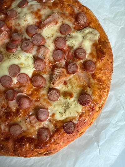 A top-down view captures a freshly baked, round pizza, partially cropped on the right side, resting on crinkled white parchment paper. The pizza features a golden-brown, visibly textured crust surrounding a base of red sauce, generously covered with melted, pale yellow cheese. Distributed across the cheese are numerous uniformly cut, circular slices of reddish-brown sausage or hot dogs. Scattered among these are irregular pieces of cooked meat, possibly ham or chicken, and thin, translucent strips that appear to be cooked onion. Dried green herbs, resembling oregano, are sprinkled over the entire surface, including the cheese and toppings. The scene is located in Hazlemere, United Kingdom. No individuals, actions, or specific interactions are depicted.