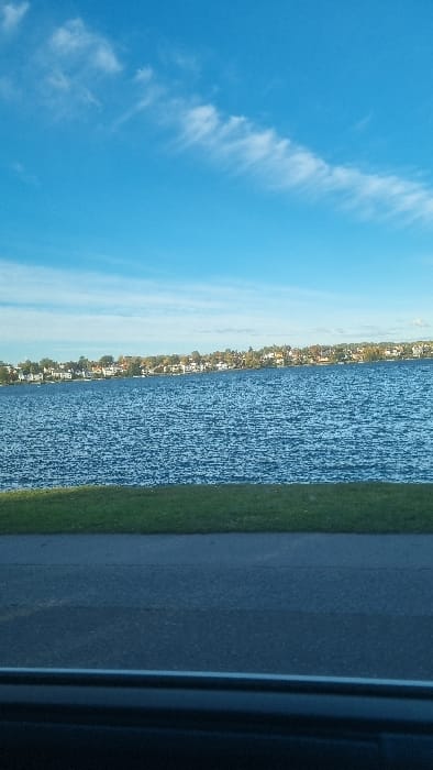 This serene landscape photograph captures a bright daytime scene over a body of water in Västervik, Sweden. The upper portion of the image features a clear, vibrant blue sky adorned with scattered, wispy white clouds that stretch horizontally across the frame.
Below the sky lies a vast expanse of deep blue water, likely a lake or a bay, its surface gently rippled, reflecting the ambient light. On the far horizon, a distant shoreline is visible, densely populated with numerous houses and buildings nestled among lush green trees, suggesting a residential area or a charming town along the water's edge.
In the foreground, closer to the viewer, a strip of well-maintained green grass borders the water, creating a natural divide. Adjacent to the grass, a dark, paved surface, possibly a road or pathway, extends across the lower part of the image. A dark, vertical structural element, likely a window frame or part of a building, partially obscures the rightmost edge of the scene, suggesting the photo was taken from an enclosed space or through an opening.
No people or specific activities are visible, contributing to the tranquil atmosphere of the scene. The clear skies and bright lighting suggest pleasant weather during the day, possibly mid-morning or afternoon. The overall impression is one of peaceful observation of a scenic waterside environment in a coastal or lakeside town. No visible text is present in the image.