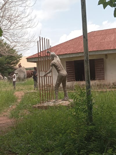 The scene depicts an outdoor art installation or sculpture garden in Eka, Nigeria. Dominating the view is a weathered, anthropomorphic sculpture appearing to leap over a metal railing. Behind the sculpture is a building with a red, corrugated metal roof and closed, wooden shutters. Several other sculptures and artworks are discernible further in the background, some appearing to depict human figures. Two individuals are present near the other sculptures, though their actions are unclear. The surrounding environment is overgrown with grass and foliage, suggesting a natural or semi-natural setting. A bare tree is visible in the foreground.