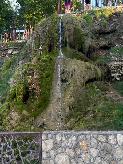 The image shows a moss-covered rock formation with water trickling down its surface, possibly a small waterfall. It is located in Krasny Klyuch, Russia. Adjacent to the rock formation is a stone wall and a decorative metal grating. Some people are standing near a metal railing at the top of the rock formation. Trees are visible in the background.