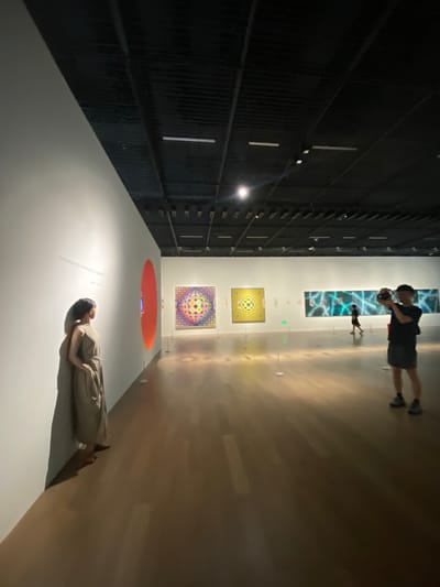 An indoor art gallery scene in the Jinniu District, China, features several individuals observing and interacting with artworks. In the foreground, a woman in a long, olive-green dress stands with her back to the camera, facing a large, bright orange circular artwork on a white wall. Her posture suggests contemplation. Further into the gallery, a man in a black t-shirt and shorts is visible from the waist down, holding a camera and appearing to take a photograph. In the background, another person, silhouetted and walking away from the camera, is seen against a long, horizontal artwork with blue and white abstract patterns. Two square artworks, one with a colorful geometric pattern and another with a yellow and black checkered pattern, are displayed on the walls. The gallery floor is a polished, warm-toned wood, reflecting the overhead lighting. The ceiling is a dark, industrial design with visible track lighting and ventilation systems. The overall atmosphere is one of quiet appreciation for the art.