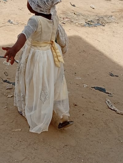 A young child, viewed from behind and slightly to the side, walks across a dirt or sandy ground in an outdoor setting in Fanisau, Nigeria. The child is dressed in a light-colored, possibly cream or pale yellow, dress. A golden sash is tied in a bow at the back of the waist. The dress has sheer white sleeves and a sheer overlay on the skirt, which is decorated with silver embroidered patterns. A white lace head covering is worn. The child's right arm is extended slightly forward with fingers splayed, suggesting movement. A dark shoe with light dot patterns is visible on the child's left foot. The ground is scattered with debris, including small pieces of plastic, fabric scraps, and organic matter. A shadow falls across the ground on the right side of the frame. The bright, clear lighting indicates it is daytime and likely sunny. No text is visible in the image.