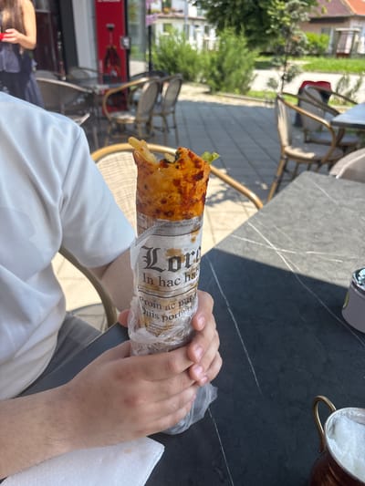 An outdoor scene in Dolni Chiflik, Bulgaria, depicts a daylight setting at a cafe or restaurant with multiple tables and chairs. In the foreground, an individual wearing a white short-sleeved shirt and grey bottoms is seated, holding a cylindrical food item with both hands. The food, appearing to be a wrap or roll, has a visible reddish-orange filling, possibly cheese or a sauce, and a piece of green lettuce protruding from the top. It is wrapped in paper resembling a newspaper, featuring "Lore" in a decorative font and Latin-like text such as "In hac ha..." and "Proin ac pu... Duis porta...". The bottom portion of the wrap is enclosed in clear plastic. A white, textured paper napkin rests beneath the individual's left hand. On the dark grey, veined tabletop to the right, a small copper-colored pot with a brass handle contains a white, frothy substance.
In the mid-ground, a woman with long dark hair, wearing a dark blue dress with ruffled details, is partially visible on the left. She appears to be holding a small red object. The background reveals additional outdoor cafe seating with chairs featuring woven backs and metal frames. Further back, a modern building with a red display or sign is visible. The distant background shows lush green trees and bushes, a paved street, and residential or commercial buildings with red-tiled roofs under bright sunlight, casting distinct shadows. The ground surface of the cafe area consists of light grey interlocking pavers.
