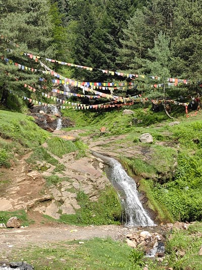 The image depicts a natural scene in Kothi, India, featuring a cascading waterfall amidst lush greenery and tall pine trees. Prayer flags, in various colors like red, yellow, blue, green, and white, are strung across the trees, creating a festive and spiritual atmosphere. The waterfall flows down a rocky incline, with smaller streams branching off. The foreground shows a dirt path with scattered rocks and grass, suggesting a rural or natural setting. The overall impression is one of serene natural beauty and cultural significance, likely a popular spot for tourists and locals alike.