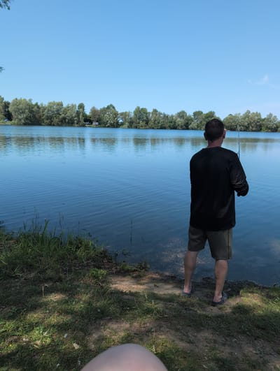 A man is fishing on the shore of a lake in Petershagen, Germany. The man is wearing a black long-sleeved shirt and olive green shorts. He is standing with his back to the camera, holding a fishing rod. The lake is calm and blue, with trees lining the opposite shore. The sky is clear and blue. The foreground of the image shows grass and dirt, with some sunlight filtering through the trees.