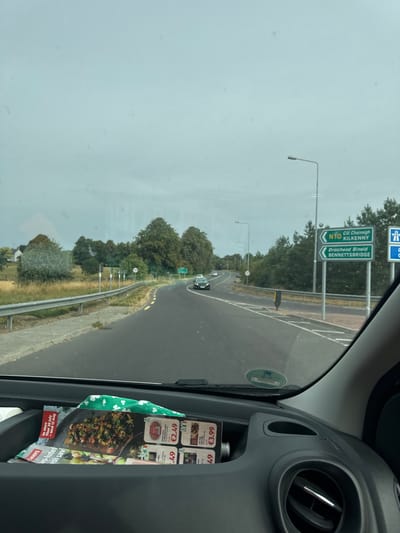 The image is a first-person view from inside a car, looking forward through the windshield. The car is on a road, with trees and fields on either side. There are road signs indicating directions to "N10 Cill Chainnigh KILKENNY" and "Draíocht Bhíde BENNETTSBRIDGE". The road appears to be in Ireland. In the foreground, on the dashboard, are some flyers and a green patterned cloth. The sky is overcast.