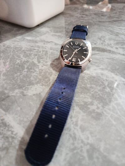 A Bulova Accutron wristwatch with a blue nylon strap lies on a grey, faux-marble surface. The watch face is dark with silver hour markers. The date window displays the number 23. Part of a white object is visible in the background. The scene is located in Bireuenmeunasahcapa, Indonesia.
