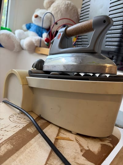 This media file shows an old iron placed on a surface with a patterned fabric. The iron has a gray handle with a cork grip and a silver metal soleplate. A black power cord extends from the iron. In the background, a portion of a white stuffed bear wearing a blue shirt is visible, along with red and blue wires. The lighting suggests it is daytime, and the overall setting appears to be indoors, possibly in a home environment. No specific activity or event is clearly depicted, but the positioning of the iron might suggest it is not currently in use. There is no visible text in the image.