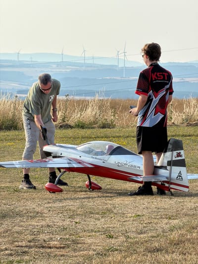 In Becherbach, Germany, a man in a khaki t-shirt and shorts, with sunglasses perched on his shirt, is bending over to adjust the propeller of a large, red and white model airplane. He is wearing black gloves. Beside him, a young man in a black and red t-shirt with the logo "KST HIGH-PERFORMANCE SERVOS" and black shorts stands holding a remote control. The model airplane, labeled "LINDINGER," is positioned on the ground. In the background, a hilly landscape is dotted with wind turbines, under a bright, hazy sky. The ground is covered in dry grass and scattered yellow flowers.